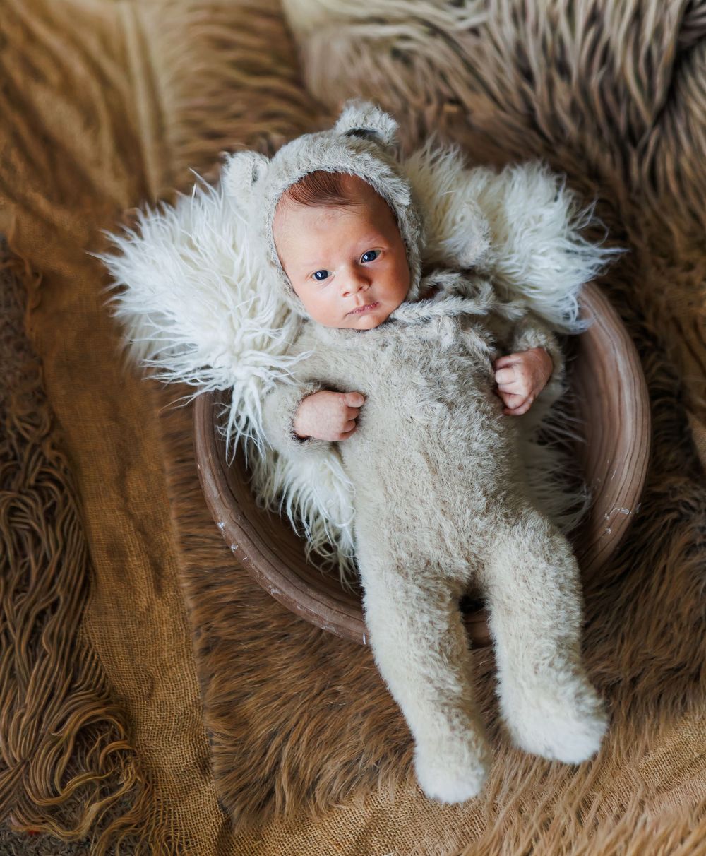 A newborn baby wearing a white knit outfit lies in a wooden bowl on a textured brown backdrop.