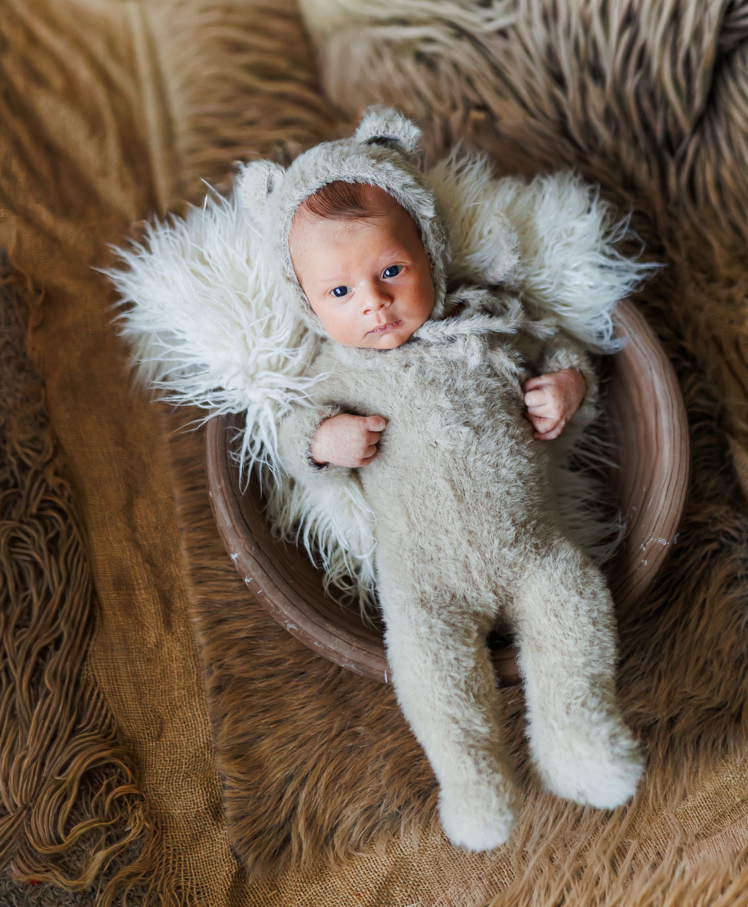 A newborn baby wearing a white knit outfit lies in a wooden bowl on a textured brown backdrop.