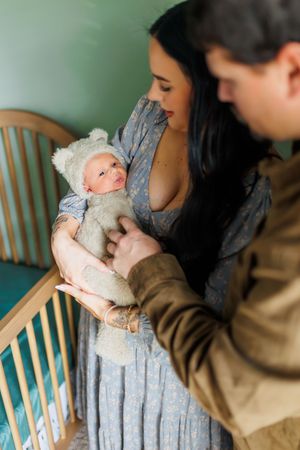Parents share tender moment with newborn baby wearing white outfit while standing next to turquoise crib.