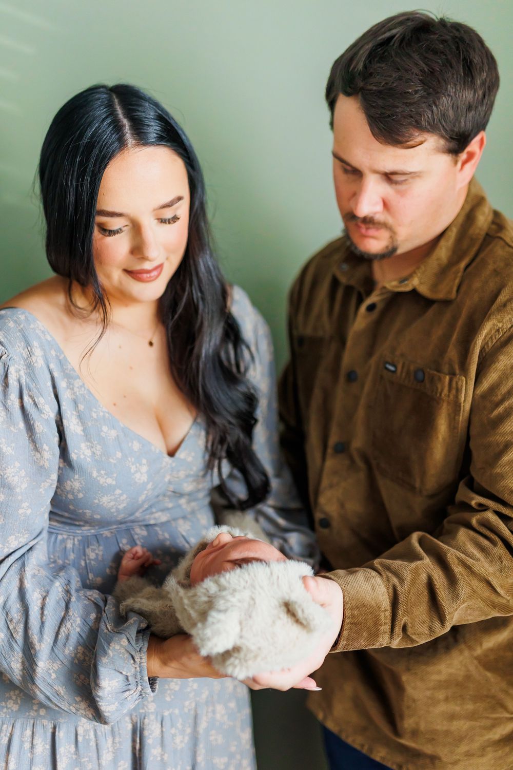 A couple in casual attire tenderly holds and gazes at their small white pet against a mint green wall.