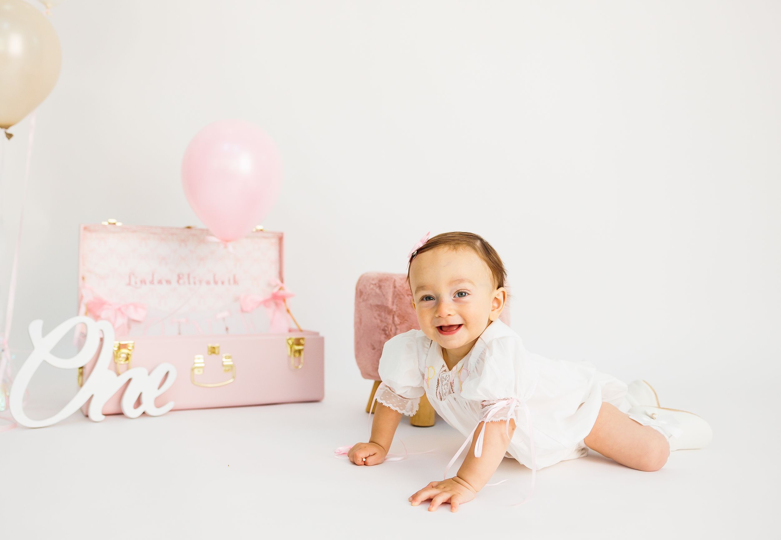 Baby girl in white outfit crawls near pink suitcase and balloon during first birthday cake smash photo session.
