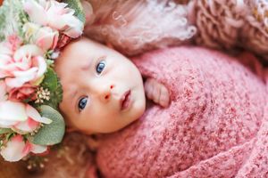 Baby wrapped in soft pink knit blanket lies next to delicate peach and pink floral arrangement.