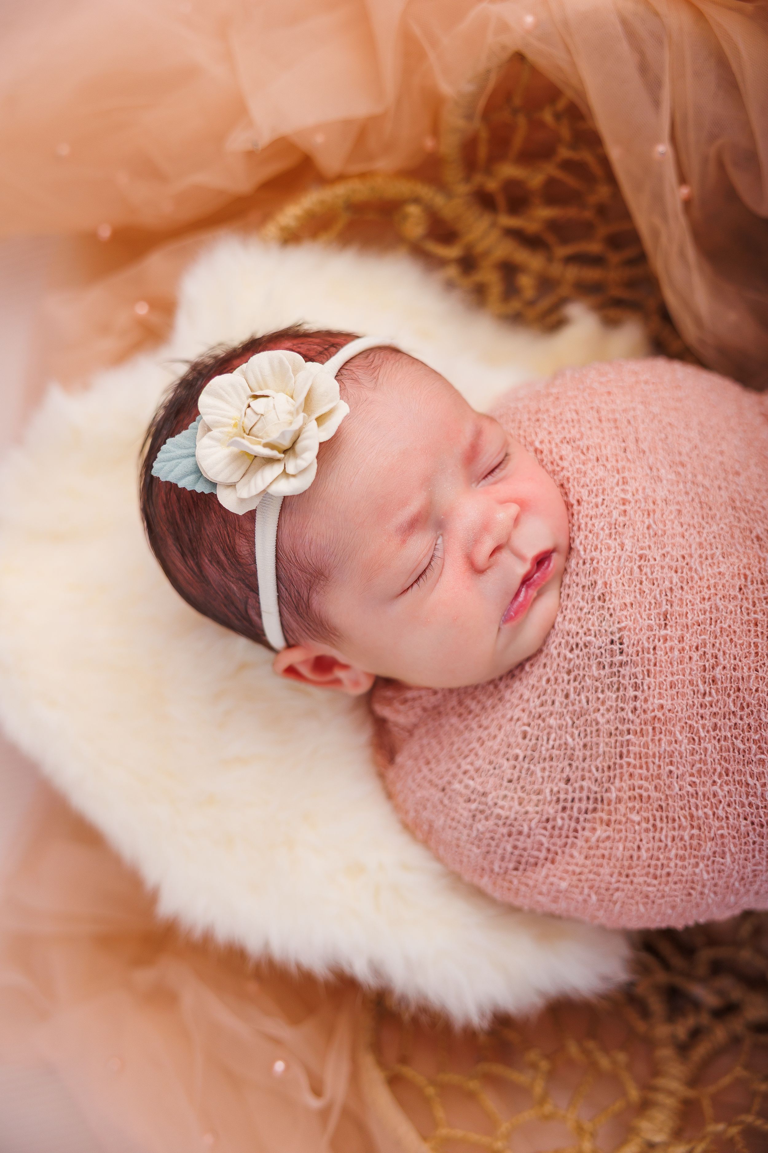 Sleeping newborn baby girl wrapped in pink knit swaddle with cream flower headband on white fur.