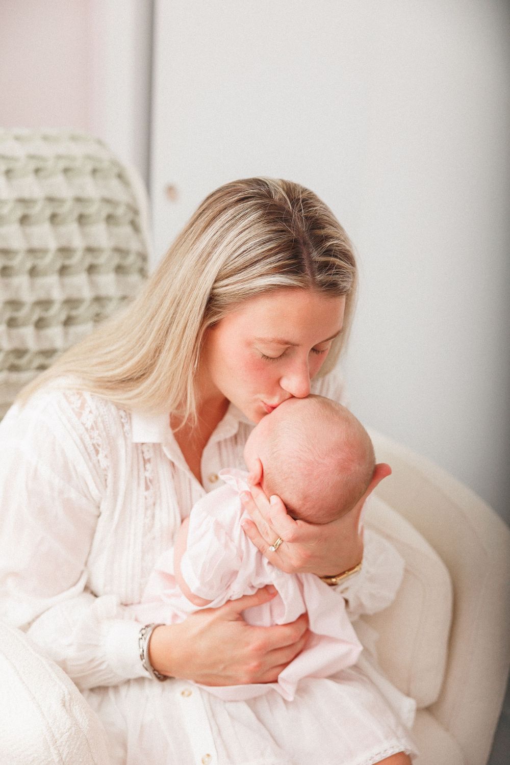 Blonde woman in white dress kissing newborn baby dressed in pink while seated in a cozy chair.