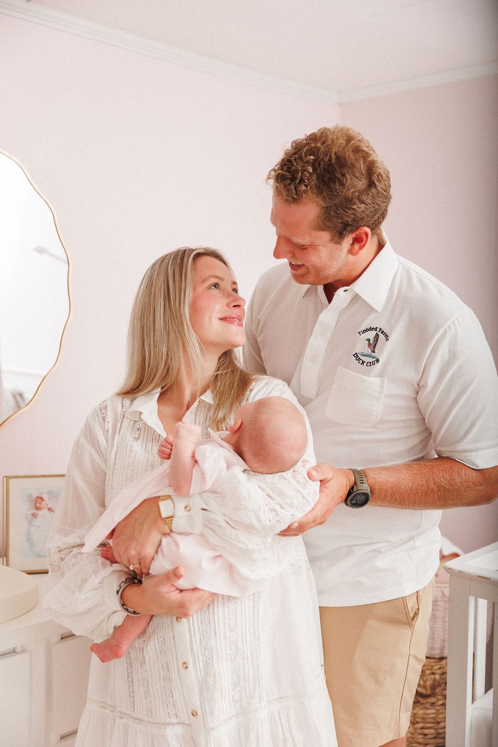 Happy couple in white outfits smiling at each other while holding their newborn baby in a pink nursery.