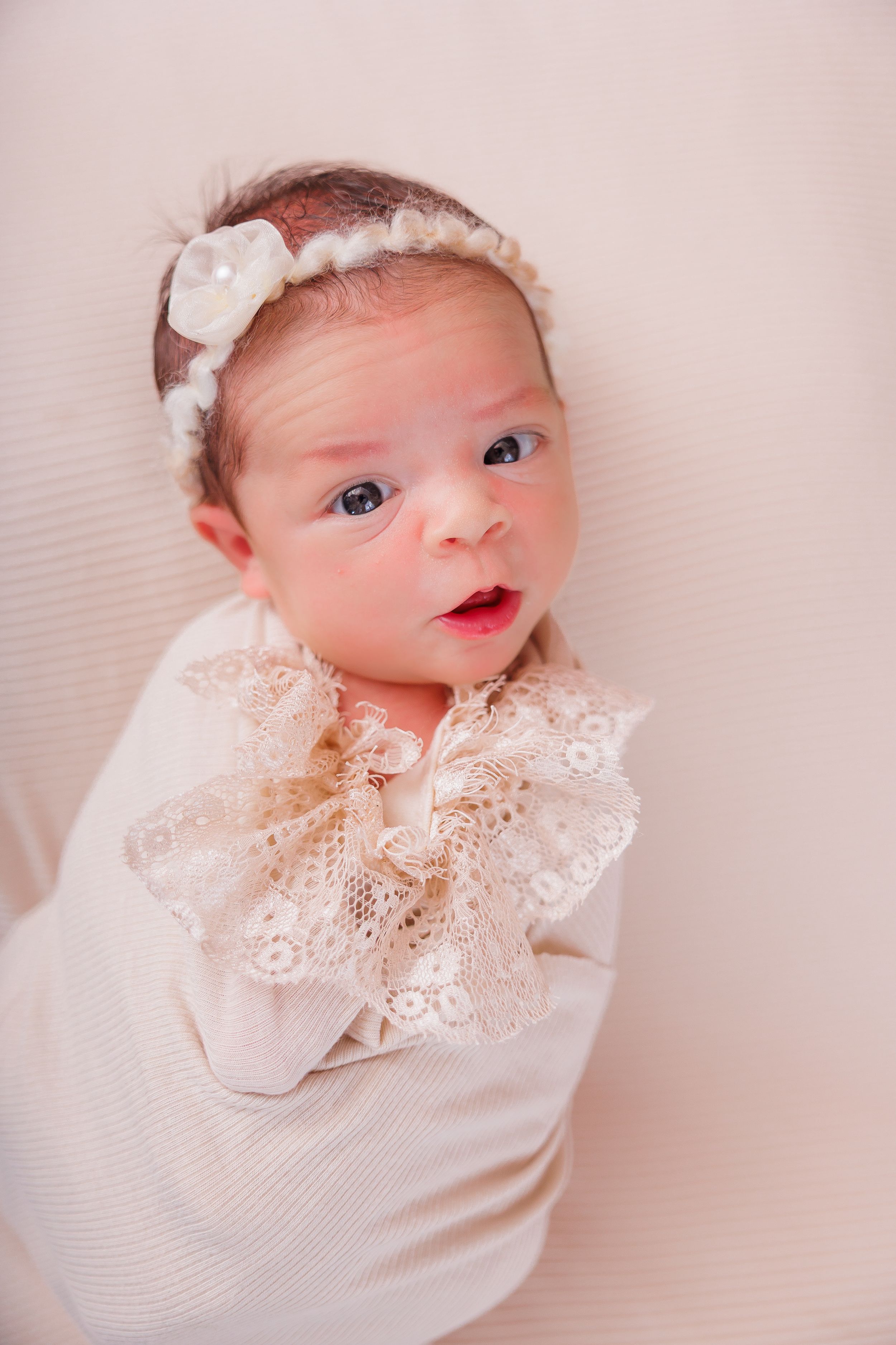 Newborn baby girl swaddled in cream wrap with lace collar and white floral headband, lying on soft background.