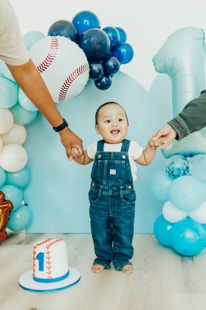 Baseball themed first birthday party setup with blue and white balloons, smash cake, and baby in denim overalls.