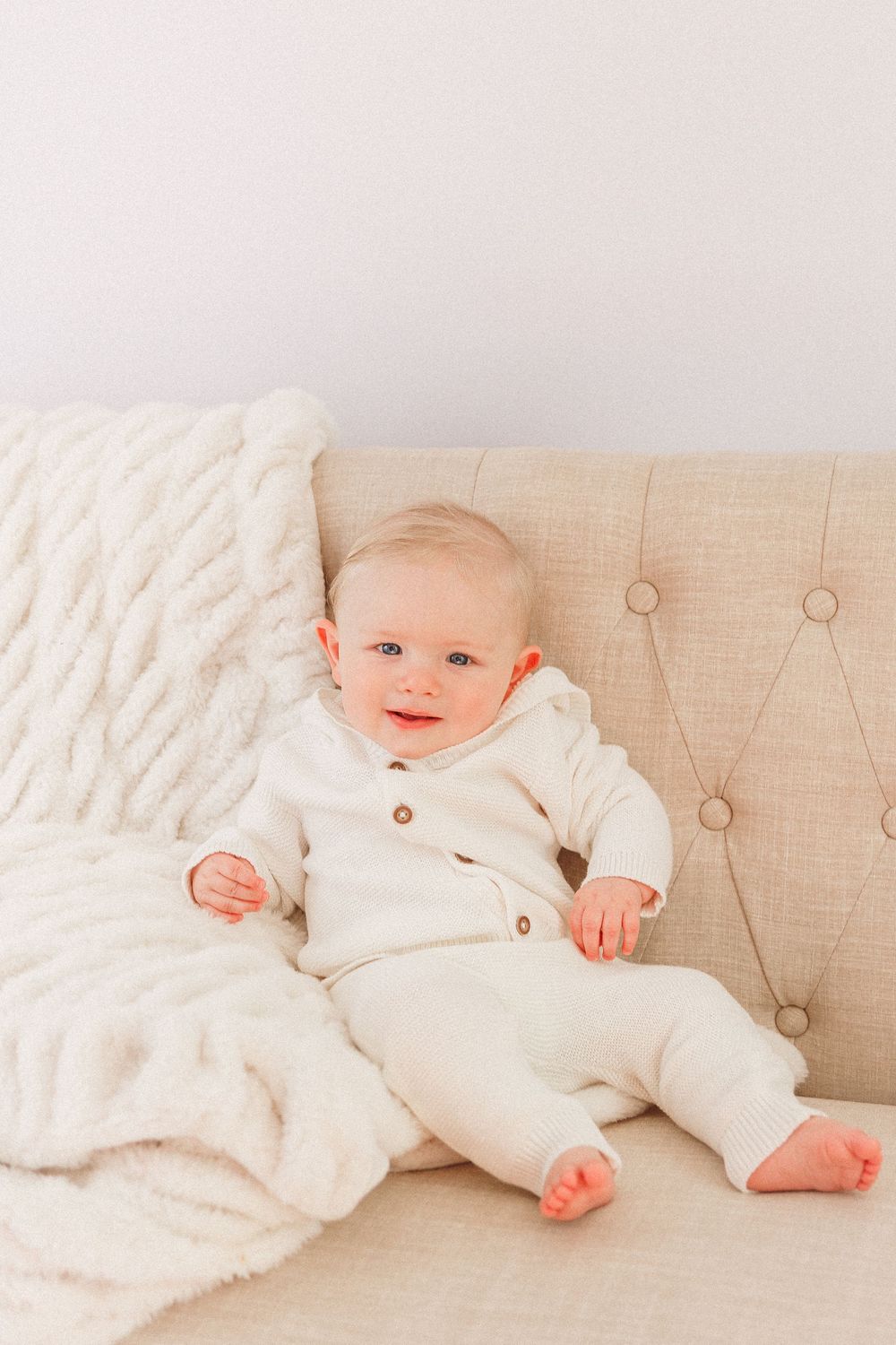 Smiling baby in white outfit sitting on beige tufted sofa with fluffy cream blanket.