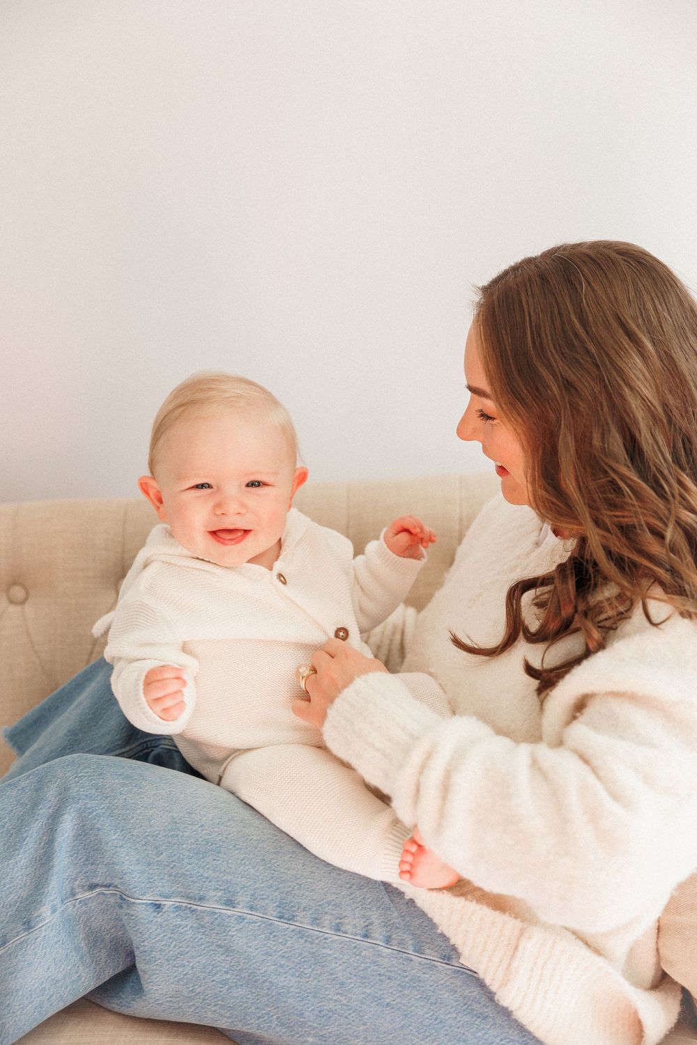 A smiling baby in white onesie sits on a couch with a brunette woman in a cream sweater and jeans.