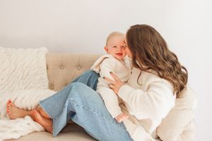 A young mother in a white sweater and jeans sits on a couch, lovingly holding and smiling at her happy baby.