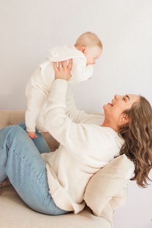 A joyful mother in a white sweater and jeans lifts her smiling baby overhead while relaxing on a beige sofa.