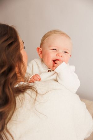 A smiling baby in a white sweater being held by a brunette mother against a light gray background.