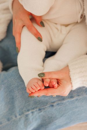 A mother with sage green nails gently holds her newborn baby's tiny feet while dressed in white knit and blue jeans.