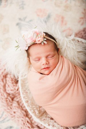 Sleeping newborn baby girl swaddled in pink wrap with floral headband in a basket with fluffy white fur.