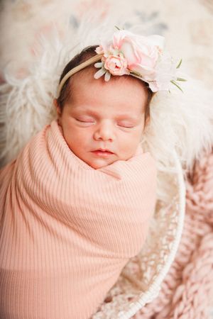 Sleeping newborn baby girl swaddled in pink wrap wearing floral headband, nestled in fluffy white basket.