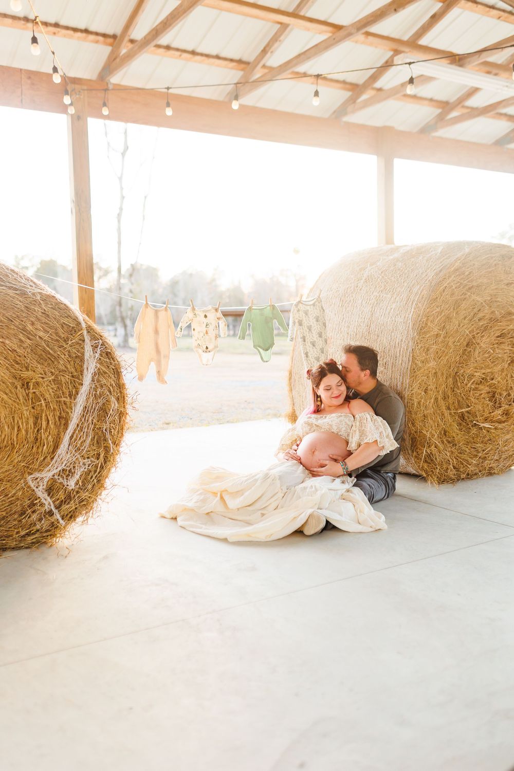 A couple in formal attire lies together on a concrete floor next to large round hay bales in a rustic barn setting.
