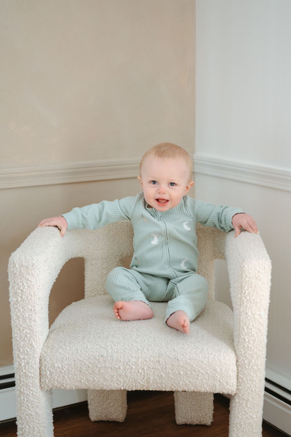 Adorable baby in mint green zip-up onesie sitting in a white boucle chair, smiling at the camera.
