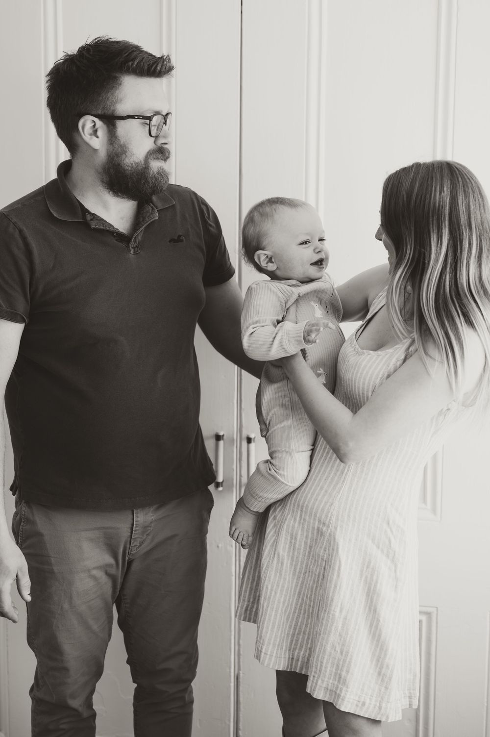 Black and white photo of a bearded father with glasses and mother holding their newborn baby together indoors.