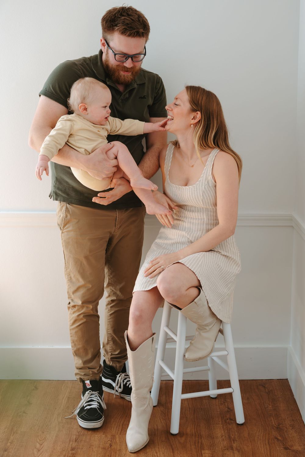 A happy young family with a bearded father holding a baby reaching for the smiling mother seated on a white stool.