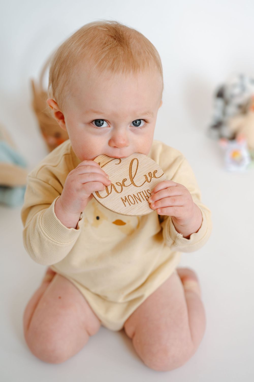 Adorable baby in yellow onesie chewing on a wooden twelve months milestone disc, sitting on white floor.