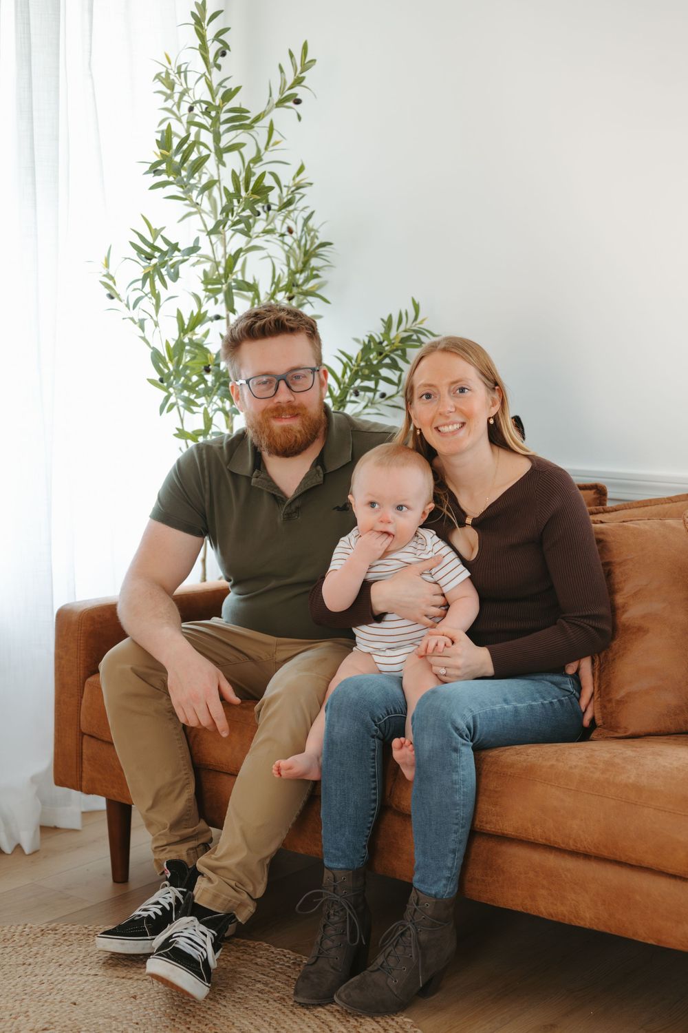 Happy young family with bearded dad, smiling mom, and baby sitting together on a brown leather couch.