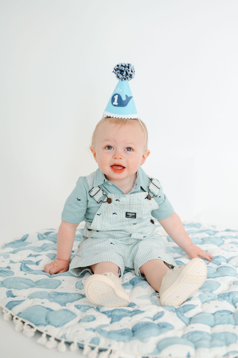 Adorable baby boy in teal overalls and birthday party hat sitting on whale-print blanket for first birthday.