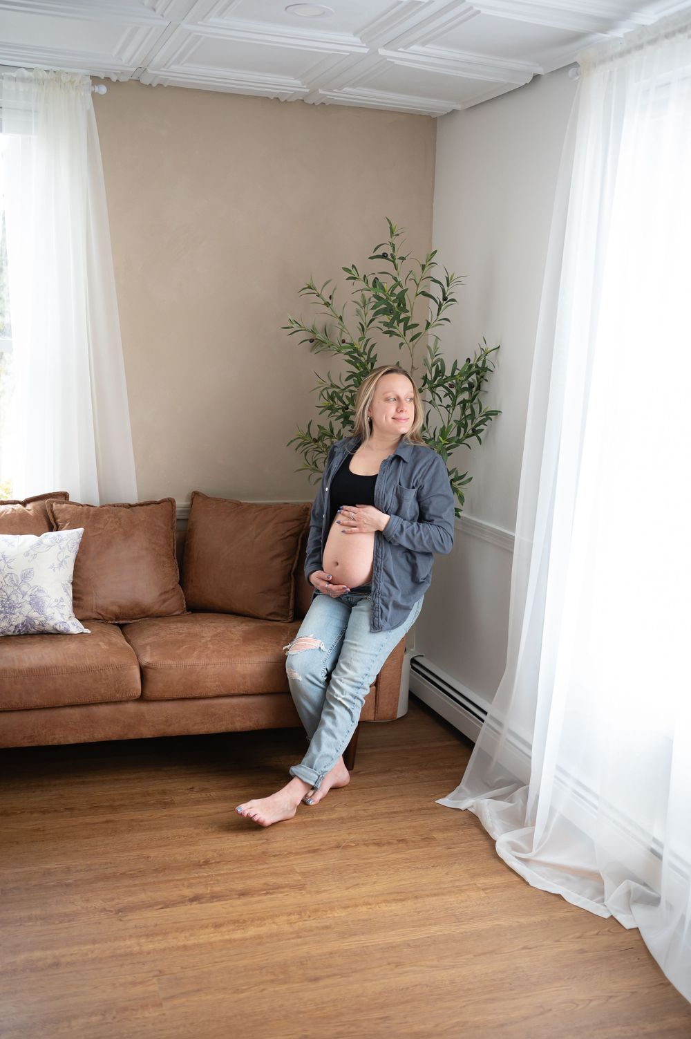 Casual interior scene showing denim-clad figure relaxing on brown leather couch with houseplant and white curtains in background.