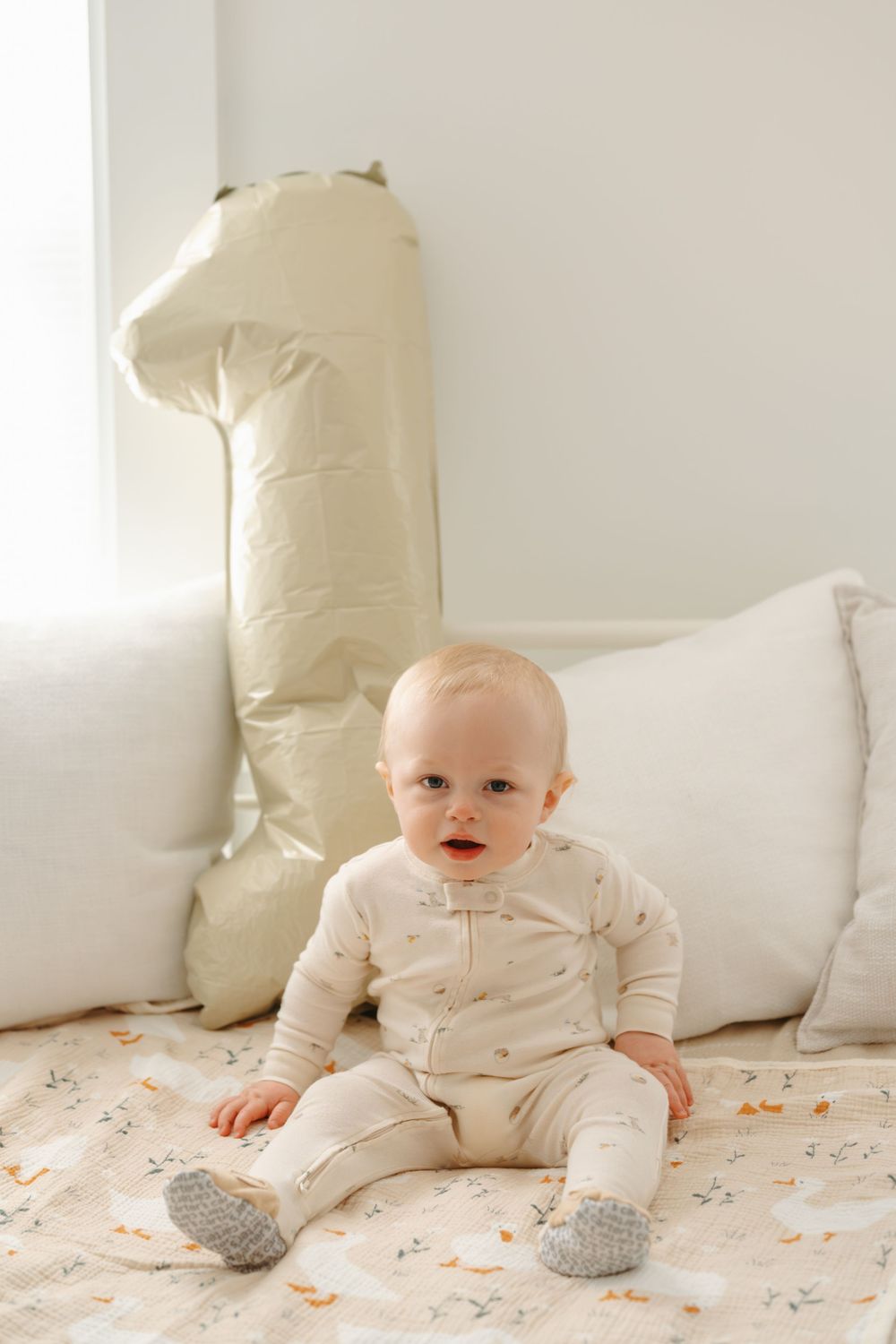 Baby in cream outfit sits on patterned blanket beside a number one balloon, celebrating their first birthday.