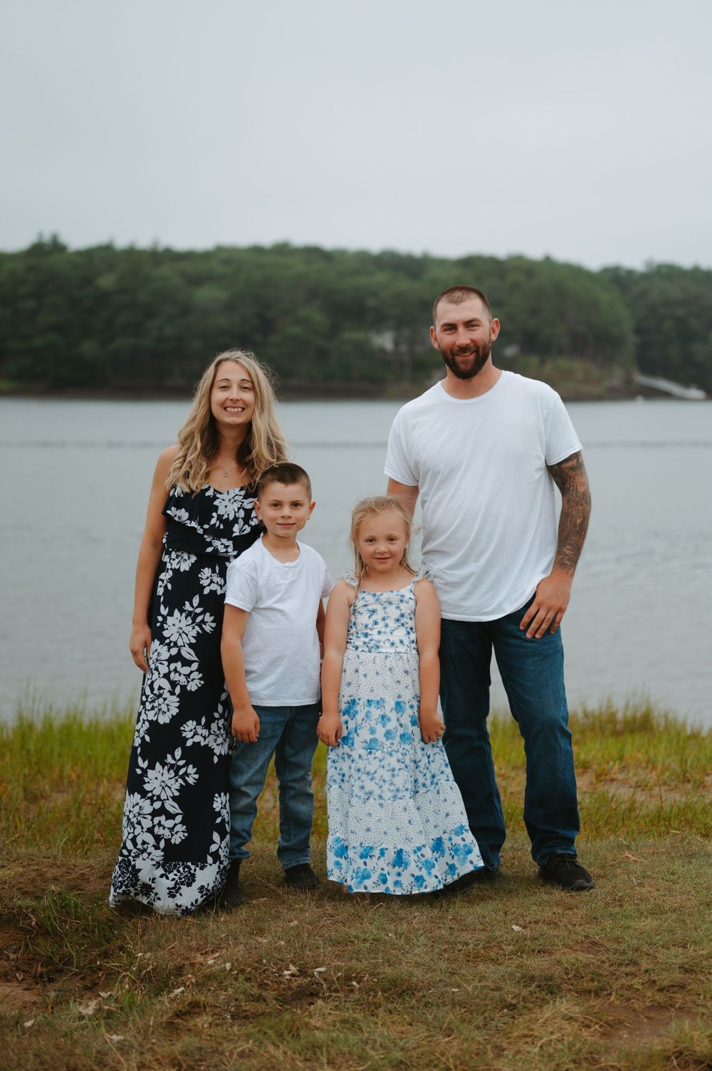 A family sits together on a grassy shore overlooking a serene waterfront on an overcast day.