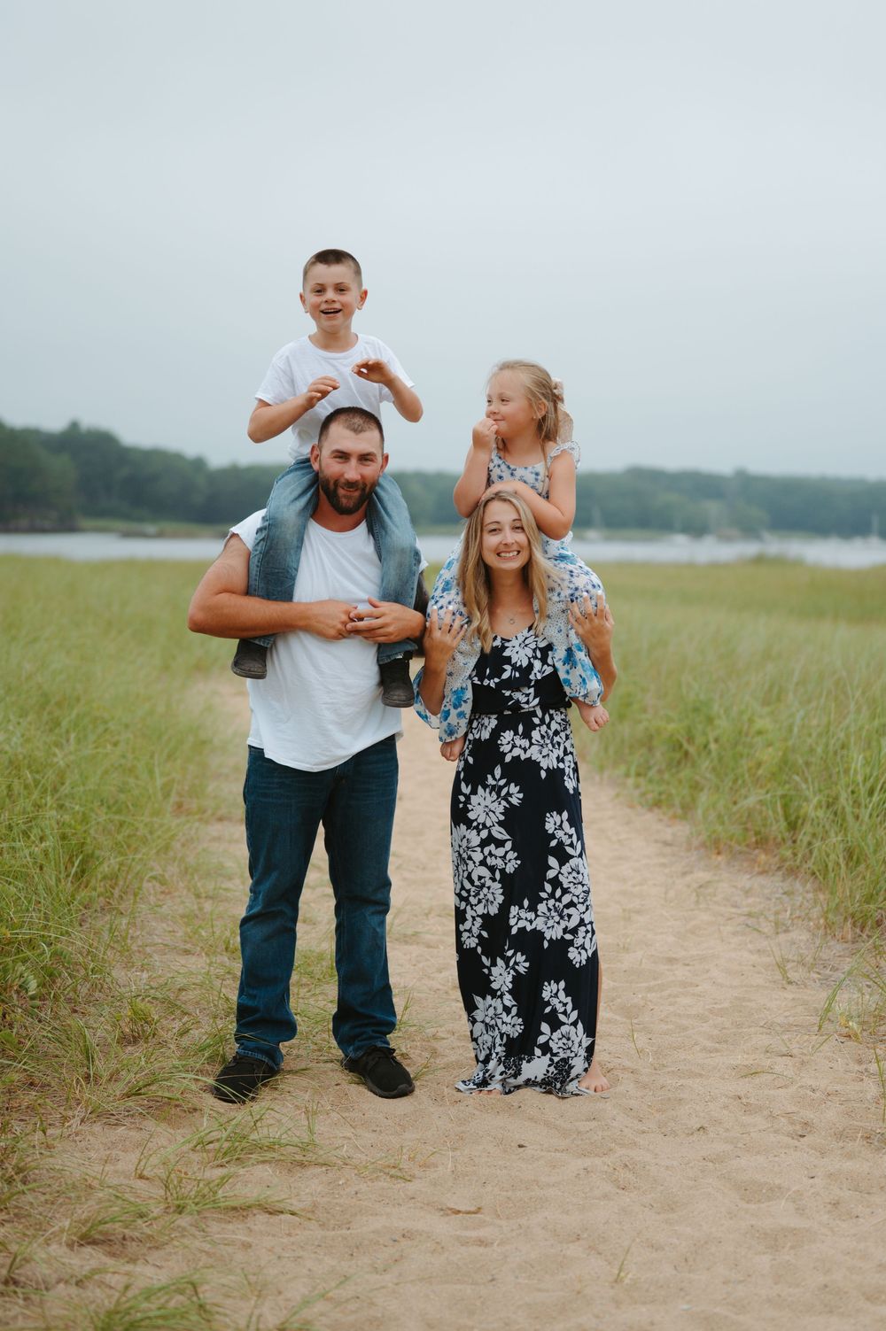 A loving family walks and plays together on a sandy path surrounded by tall grass near a lake.