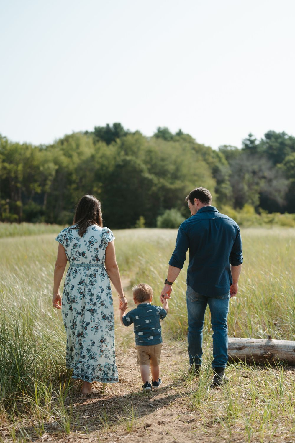 A family walks hand in hand with their toddler along a dirt path through a sunlit meadow on a summer day.