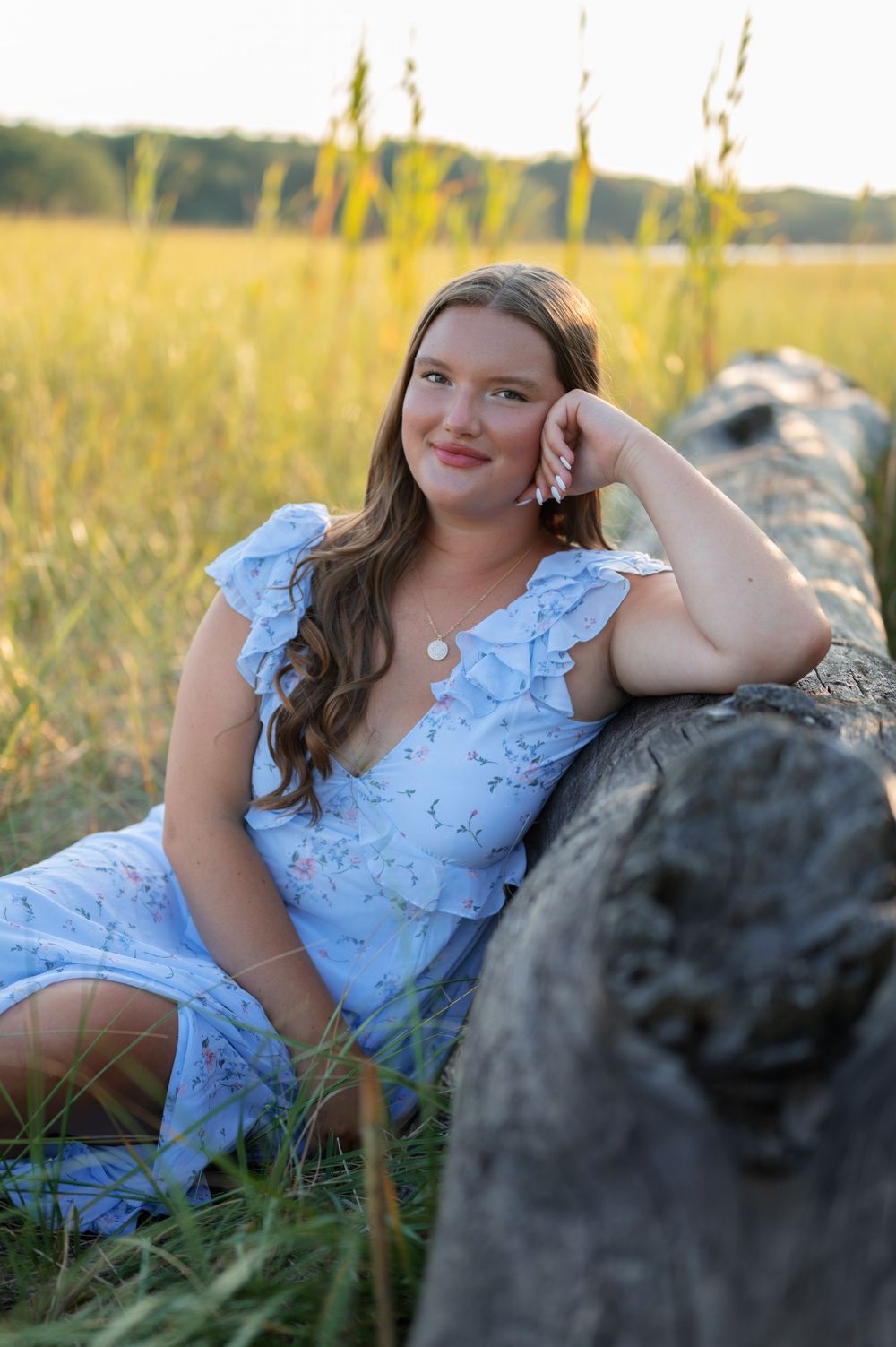 A soft blue dress stands out against tall golden grass as someone poses beside a large rock at sunset.