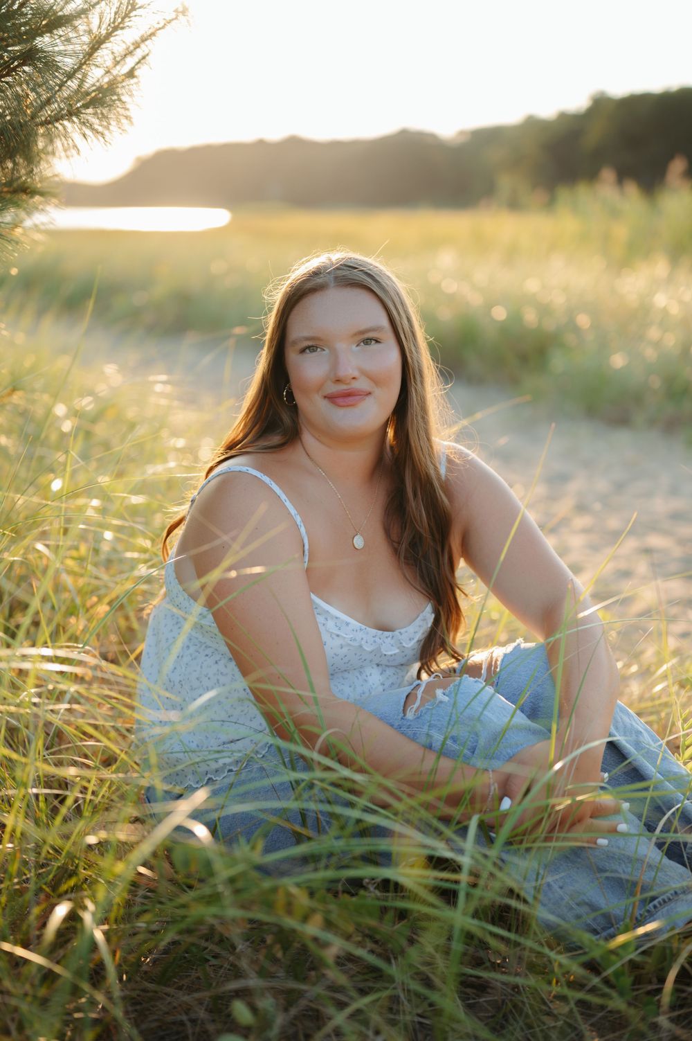 Person in a white dress sits in golden beach grass during sunset with water in background.