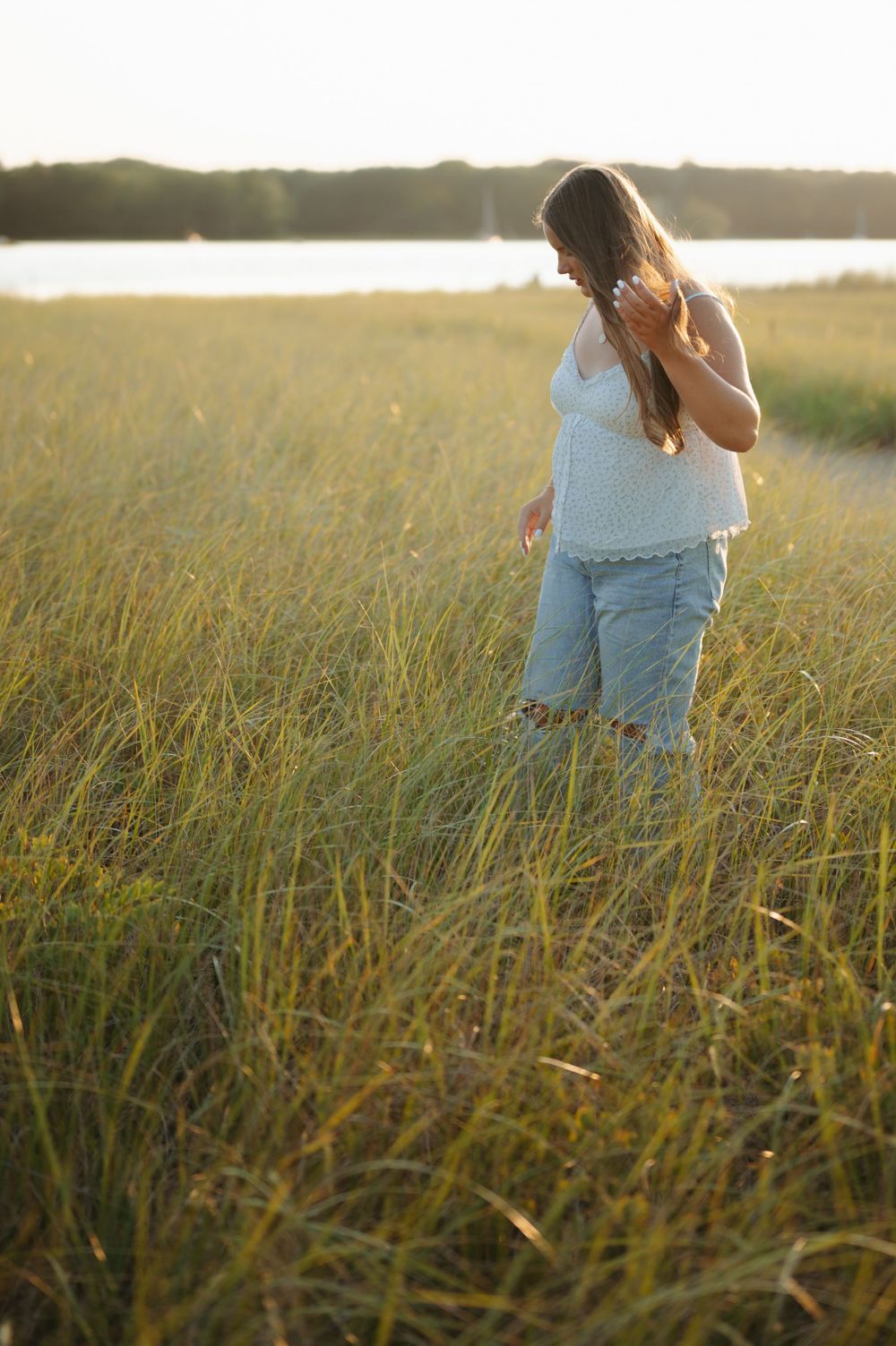 Person in a white tank top and jeans stands in a golden field at sunset with a lake in the background.