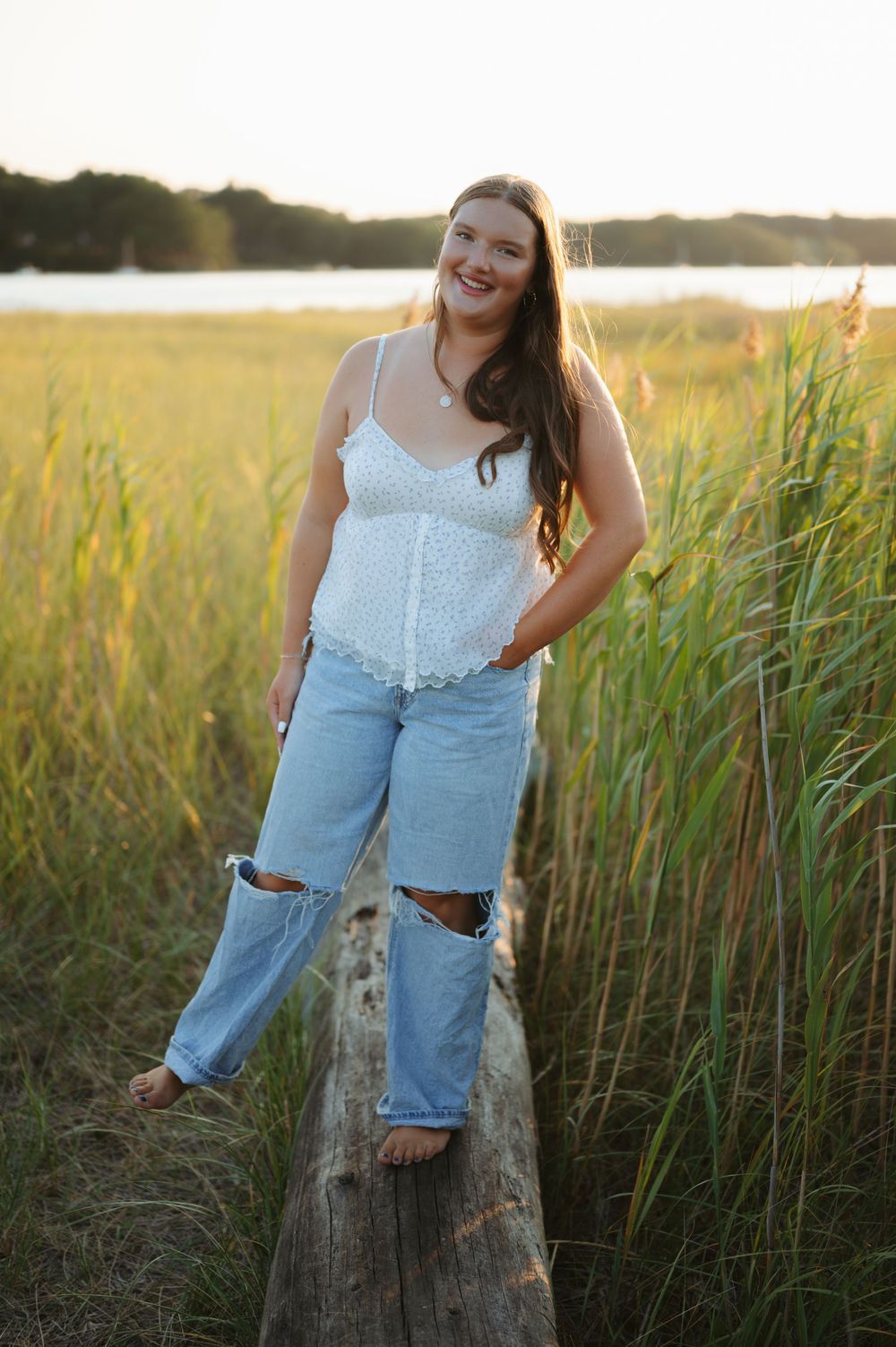 Person wearing white top and ripped jeans stands on log in coastal grass during golden hour.