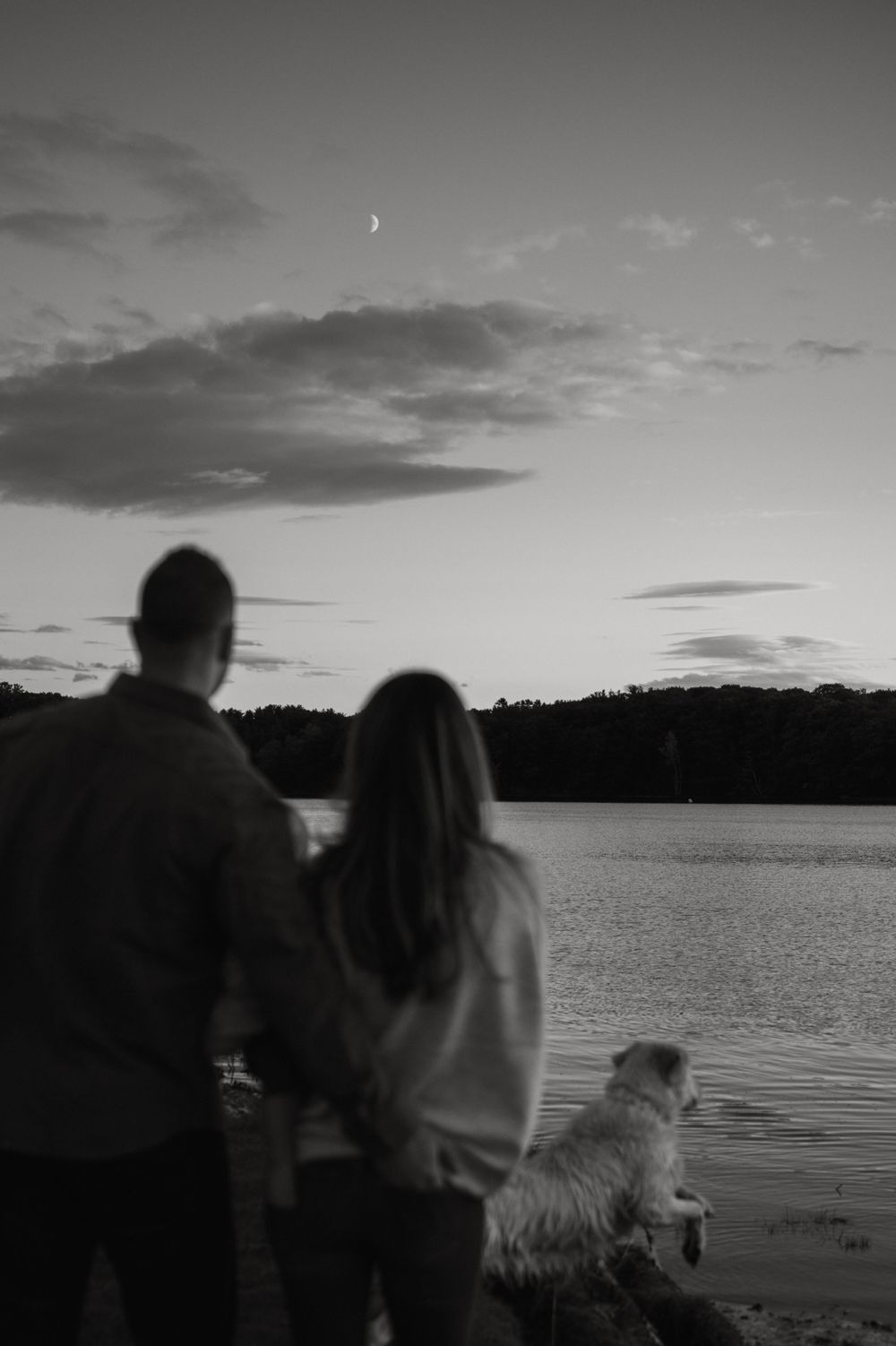 A couple and their dog watch the sunset over a lake in black and white.