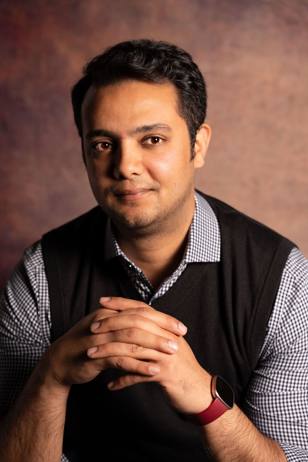 Professional portrait of a person in checkered shirt and vest against textured brown background.