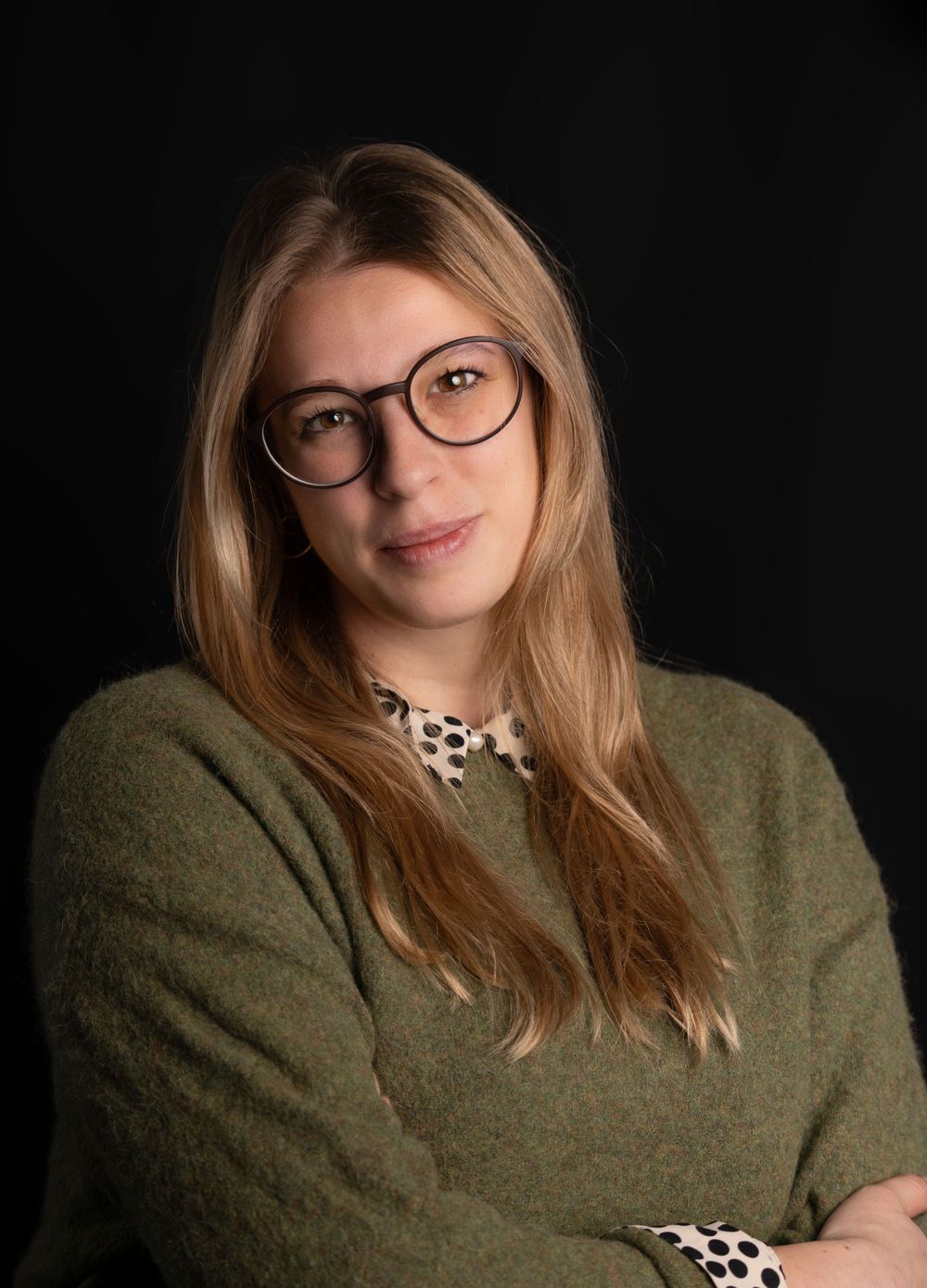 Professional portrait of a person in glasses and olive sweater against dark background with soft lighting.