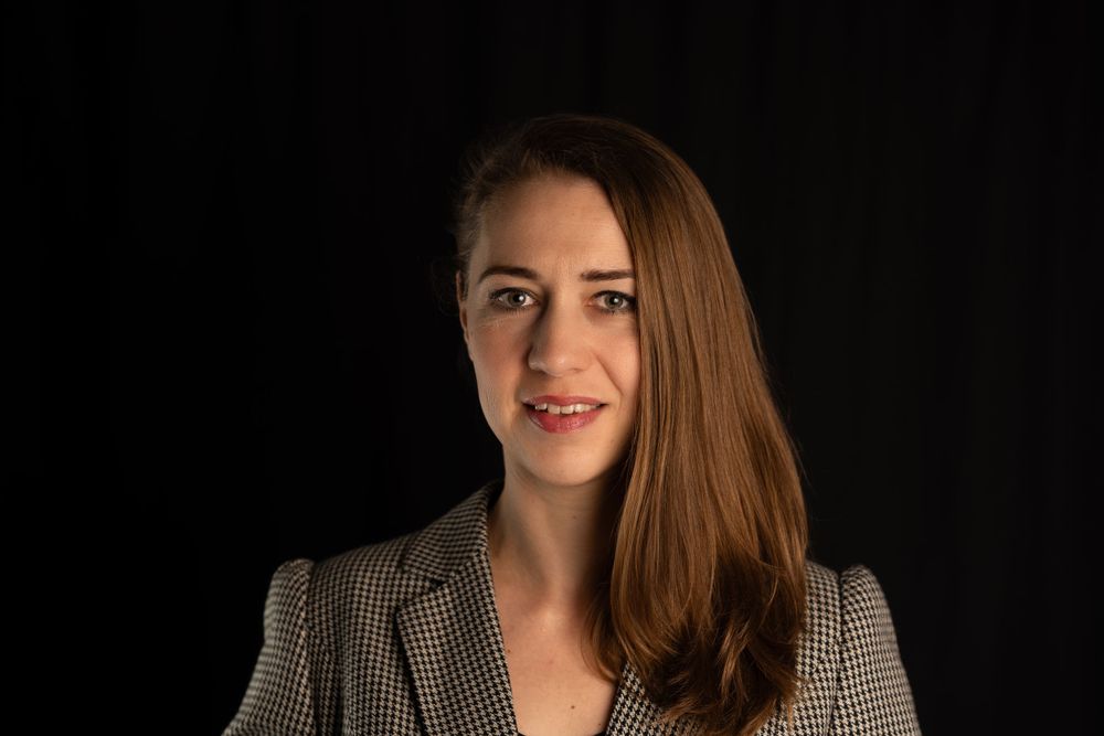 Professional headshot of person in checkered blazer against dark background.