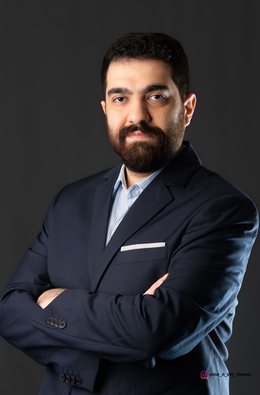 Professional portrait of a bearded man in dark suit with crossed arms against black backdrop.