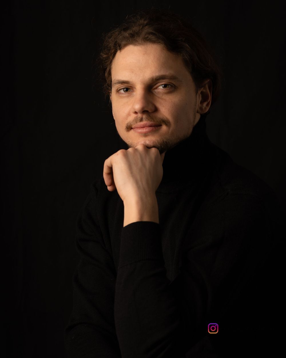 Professional portrait of a person in black turtleneck with hand on chin against dark background.