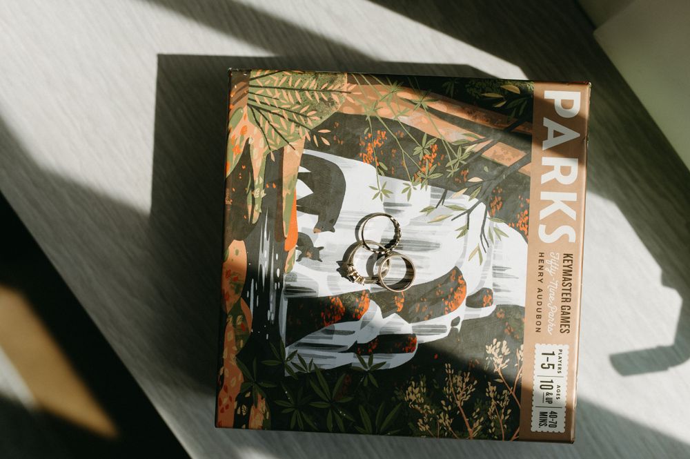 Board game box with nature-themed artwork and wedding rings in natural lighting.