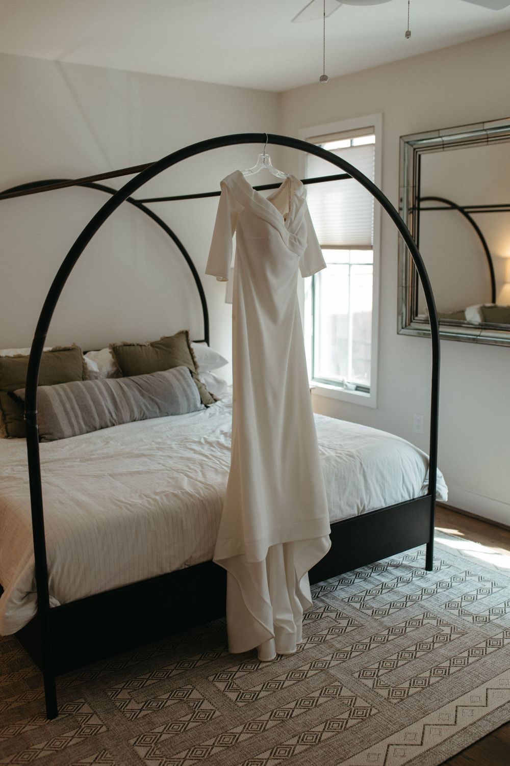 An elegant white wedding dress hangs on a black metal canopy bed frame.