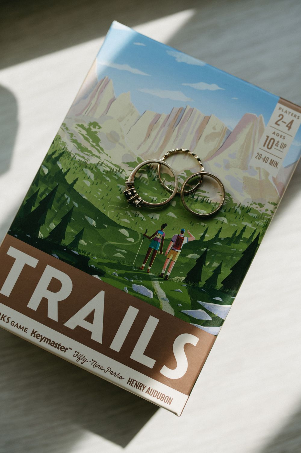Wedding rings displayed on a board game box titled Trails featuring mountain landscape artwork.