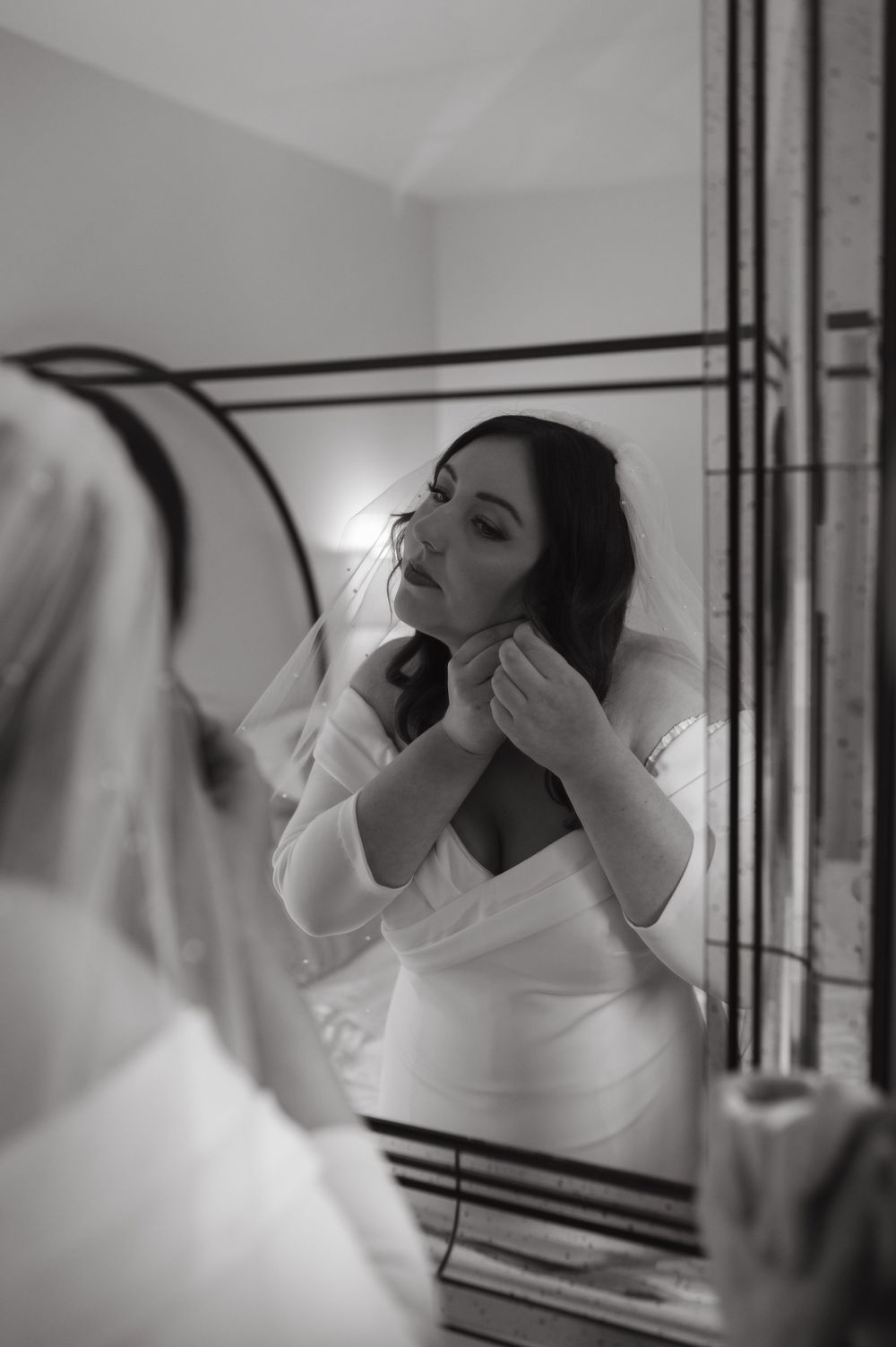 Black and white photo of a bride adjusting her jewelry in a mirror reflection.