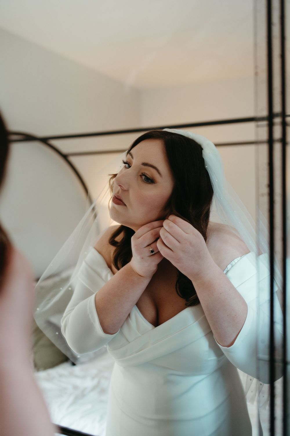 A bride adjusts her earrings while wearing an elegant white wedding dress and veil.