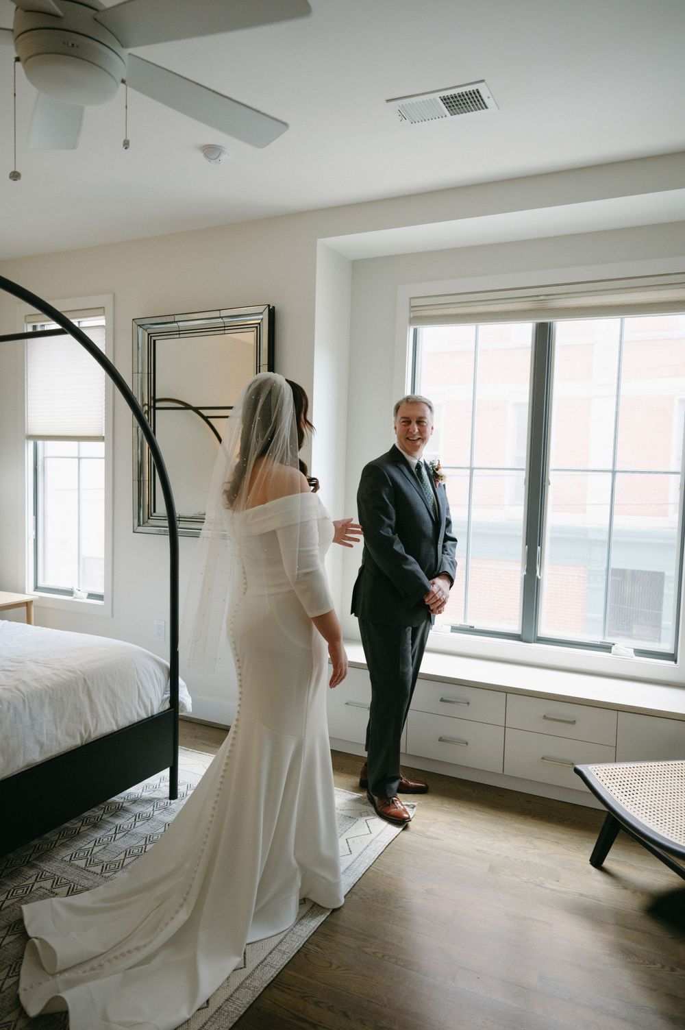 A wedding couple stands in conversation by a window in an elegant bedroom.