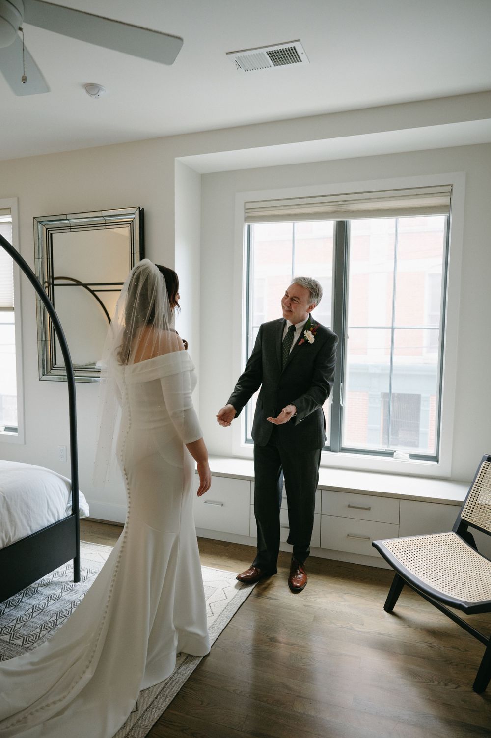 A wedding couple shares a moment in front of a window in a modern bedroom setting.