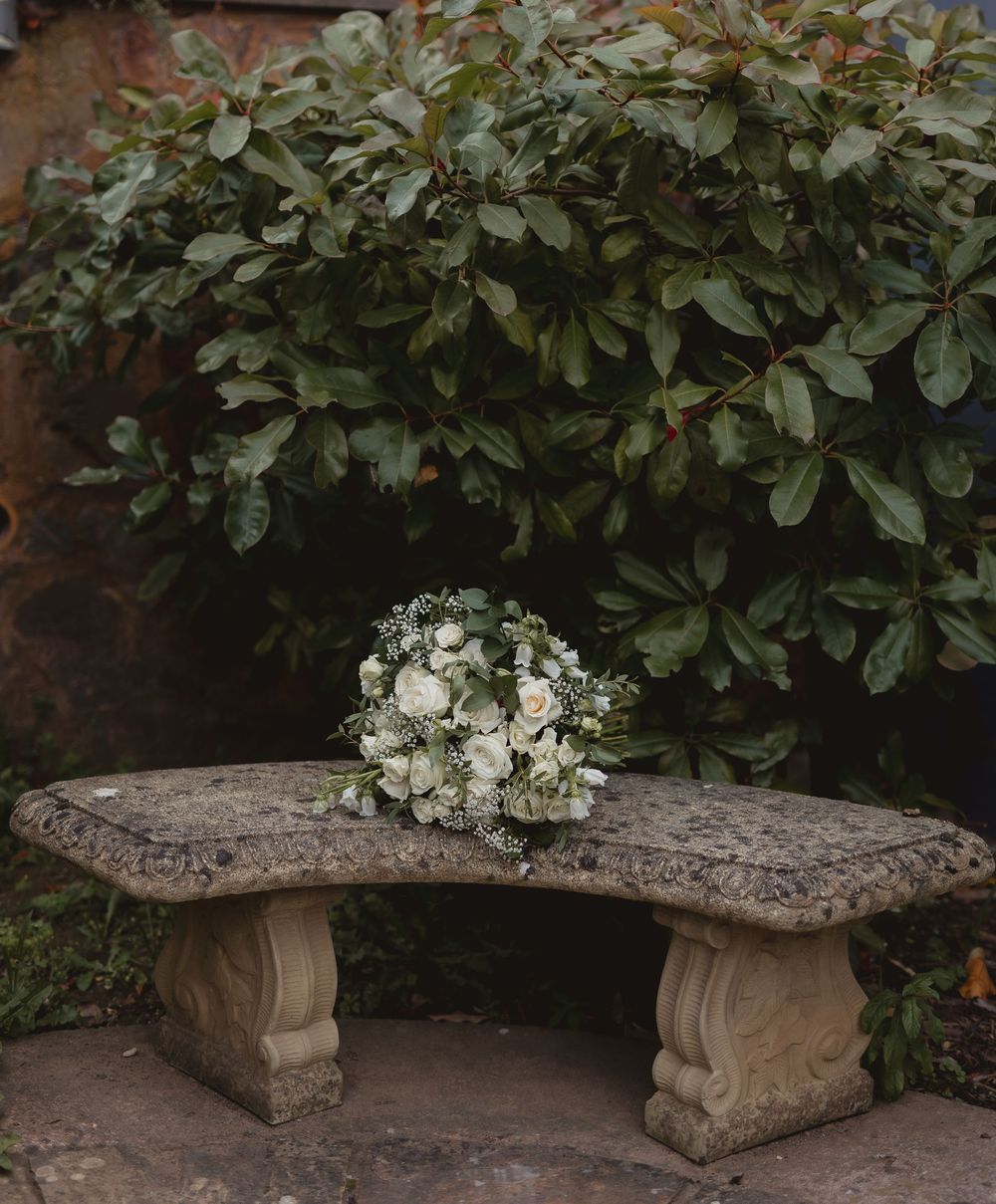 Elegant curved stone garden bench adorned with white floral bouquet arrangement against lush green foliage background.
