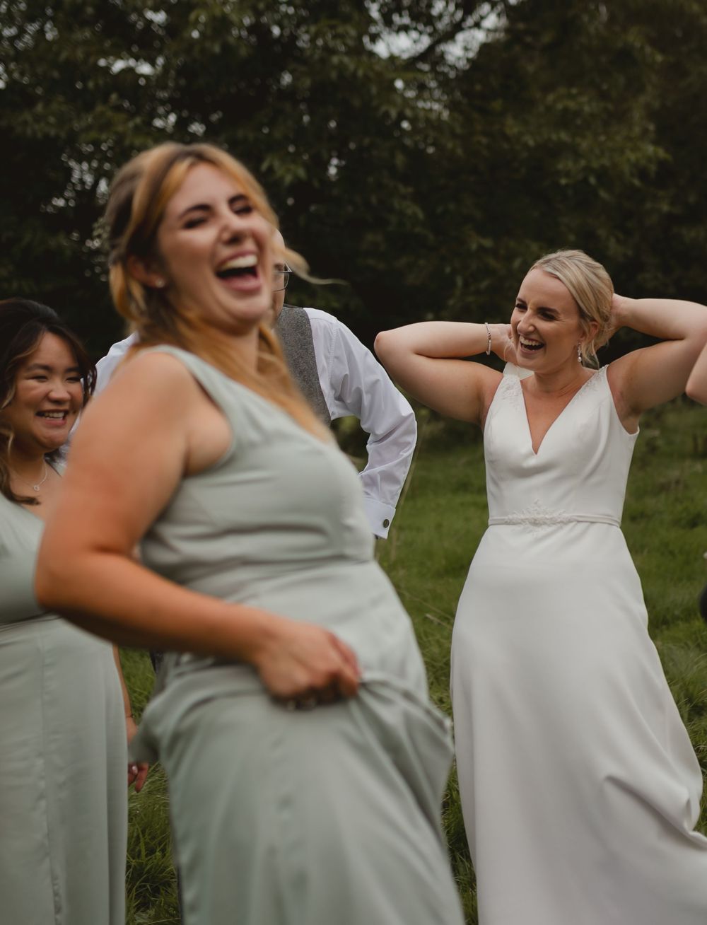 Group of bridesmaids in matching sage green dresses laughing and sharing a candid moment outdoors.